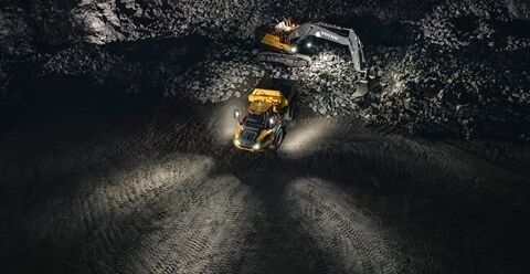 一辆沃尔沃铰接式卡车正在夜间的采石场上作业，负责装载的是一台沃尔沃挖掘机。机器上的工作灯照亮了四周。