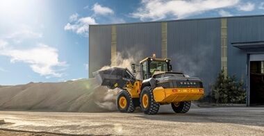 Side view of Volvo L120H Electric lifting up sand with its front-mounted bucket in front of a depot building.