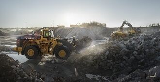 Side view of a L350H wheel loader with full bucket of rocks with Volvo excavators and haulers in the background.